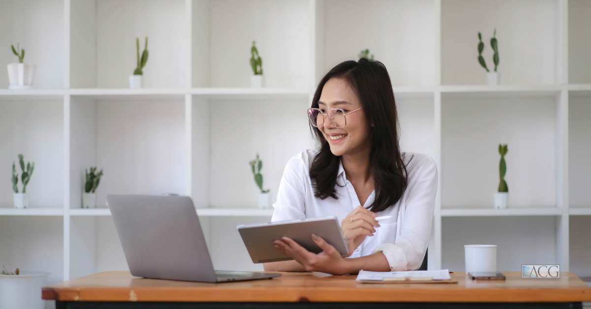 A smiling woman is working on a laptop at a desk in a modern office space.