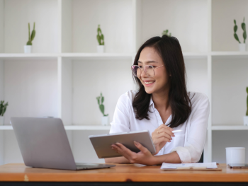 A smiling woman is working on a laptop at a desk in a modern office space.