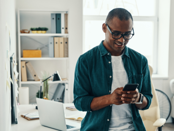 A young man looks at his mobile phone while standing in his home office.