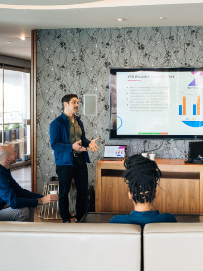 A business professional presenting growth metrics on a screen to a small group in a modern office.